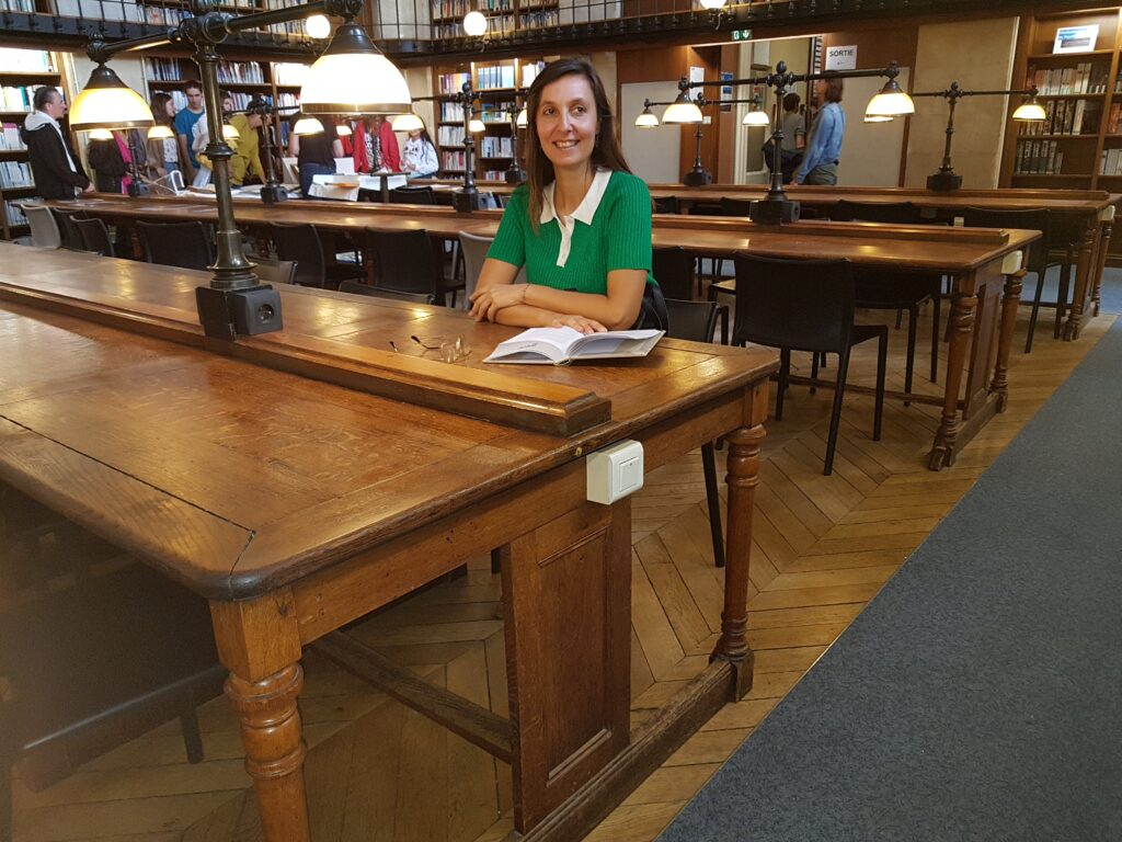 La chercheuse est assise à la table d'une bibliothèque. Ces tables sont disposés en grandes enfilades avec des lampes faisant penser à des lanternes. En arrière-plan, on voit de grandes bibliothèques remplies de livre sur deux étages.