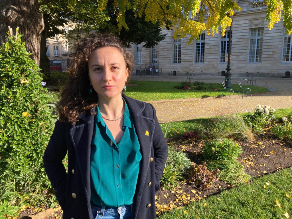 Morgane Robert est une jeune femme aux cheveux bruns et bouclés, elle se trouve dans un jardin ensoleillé. A l'arrière plan on voit aussi un bâtiment et des chaises.