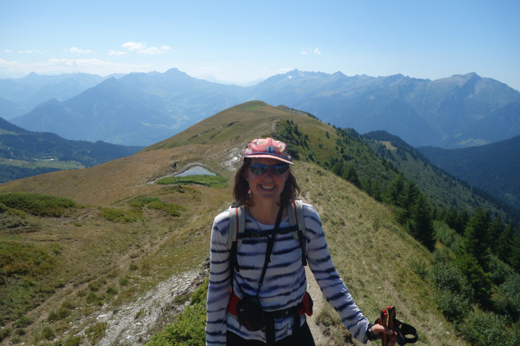 La chercheuse est en pleine randonnée, sous le soleil, caquette et lunettes de soleil. Derrière elle, de belles montagnes et quelques points d'eau.