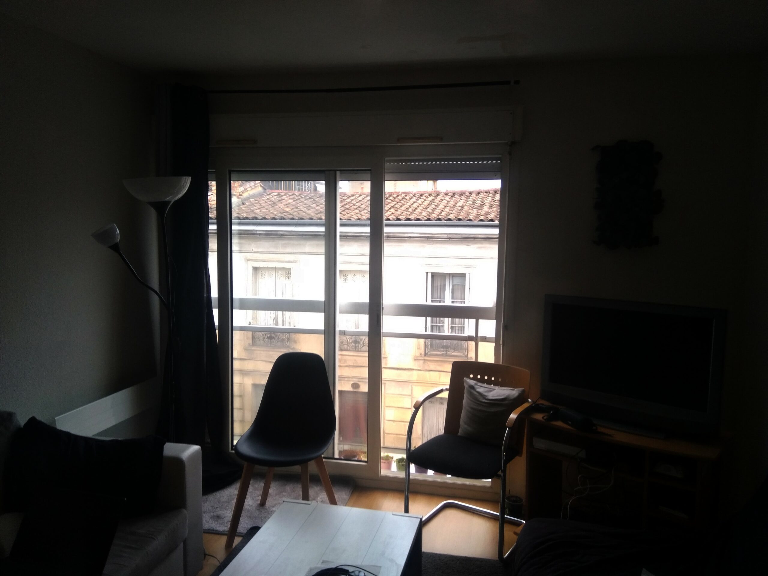 Photo d'un appartement. Seule une porte-fenêtre permet d'apporter de la lumière. L'ambiance est sombre, l'autre côté de la rue est visible.