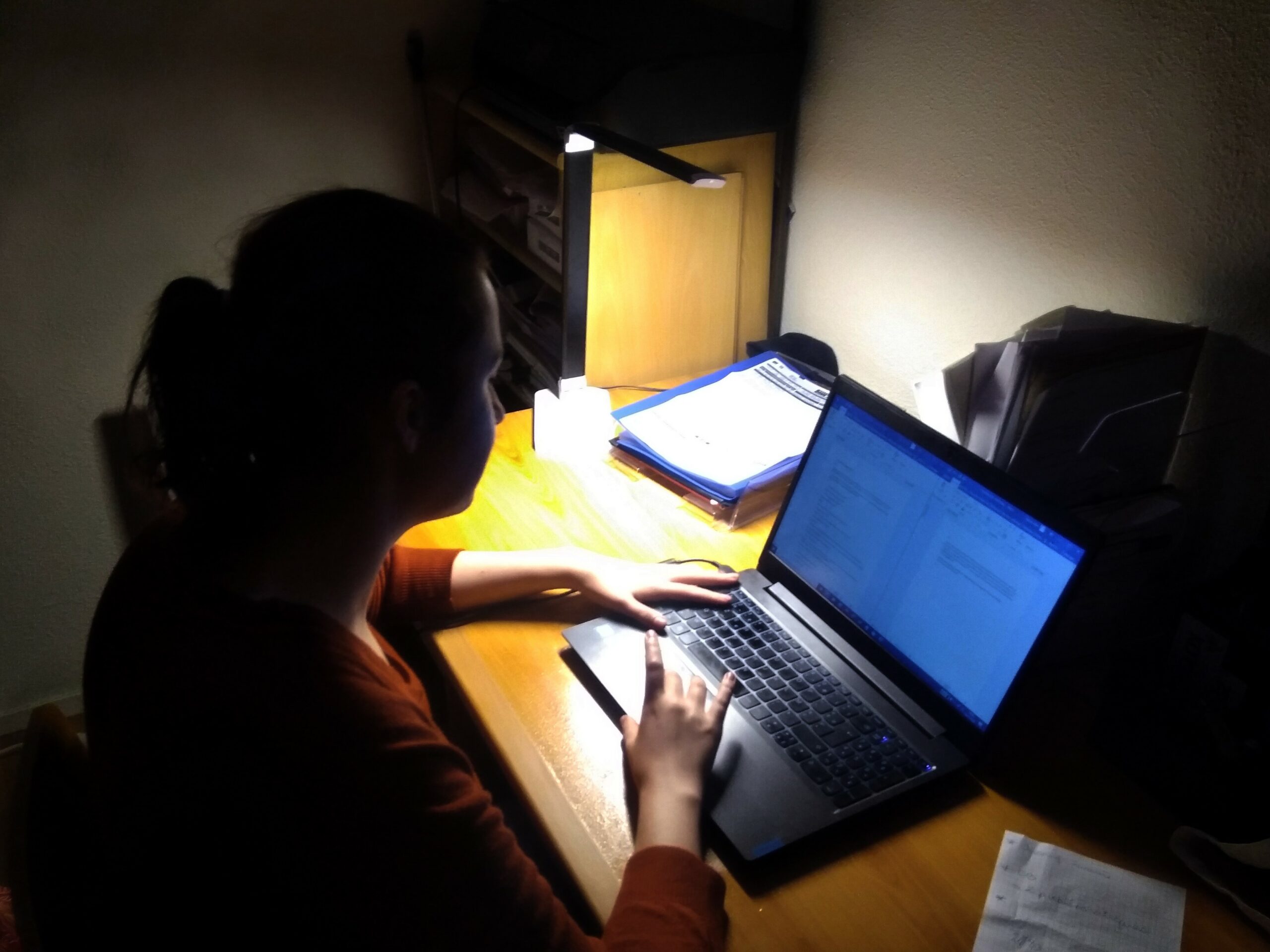 Photo d'Aurore travaillant calfeutrée dans le noir et éclairée uniquement par son écran d’ordinateur et une lampe de bureau dérisoire.