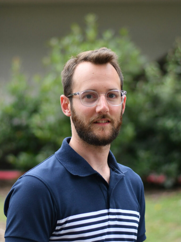 Portrait de Matthieu Guemann en plan poitrine. Il pose souriant et le regard fixé à la caméra devant un fond naturel et boisé. Il a les cheveux châtains clairs, une barbe courte de la même couleur que ses cheveux et des lunettes transparentes. Il porte un polo rayé bleu et blanc.