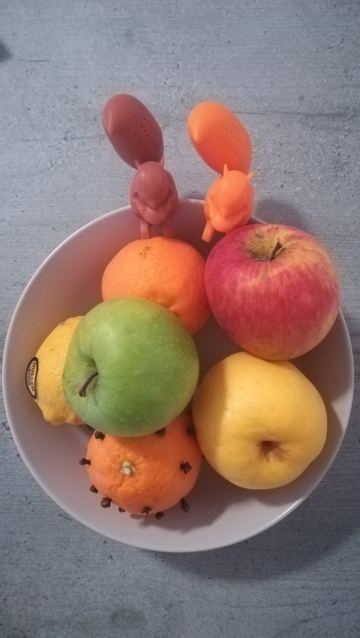 Sur une table grise, un plateau de fruits est photographié d'en haut. Celui-ci contient trois pommes : une rouge, une jaune et une verte, au-dessus. Un citron, sur la gauche, arbore une étiquette. En bas de l'image, une clémentine est percée de clous de girofles, lui conférant une apparence de hérisson. Plus haut, une autre clémentine, est lisse, elle. Sur les bords hauts du plateau, deux infuseurs à thé en forme d'écureuils sont côte à côte. Celui de gauche est marron, l'autre, à droite, orange.