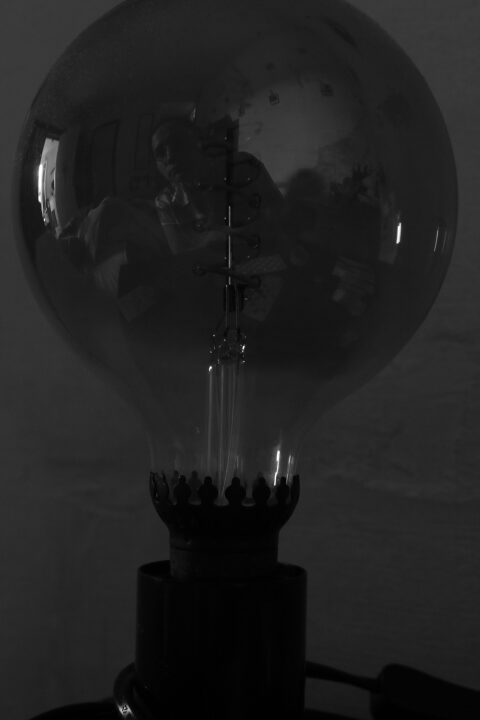 Une lampe de chevet avec une grosse ampoule éteinte, dans lequel se reflète Clémence et son appartement. L'image est en noir et blanc.