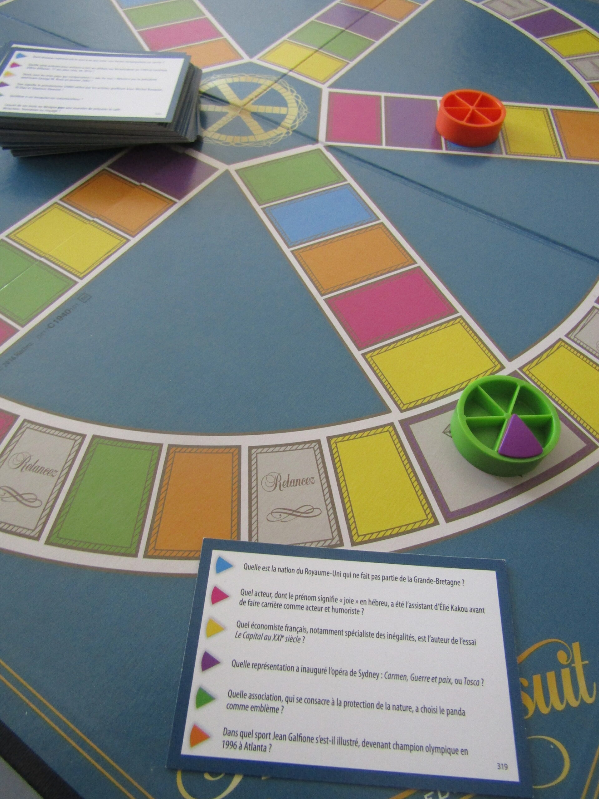 Un jeu de société Trivial Pursuit en cours de partie. On observe une carte avec des questions au premier plan suivie d'un plateau en second plan. Le plateau est composé de nombreuses cases colorées et on peut observe 2 pions sur le plateau.