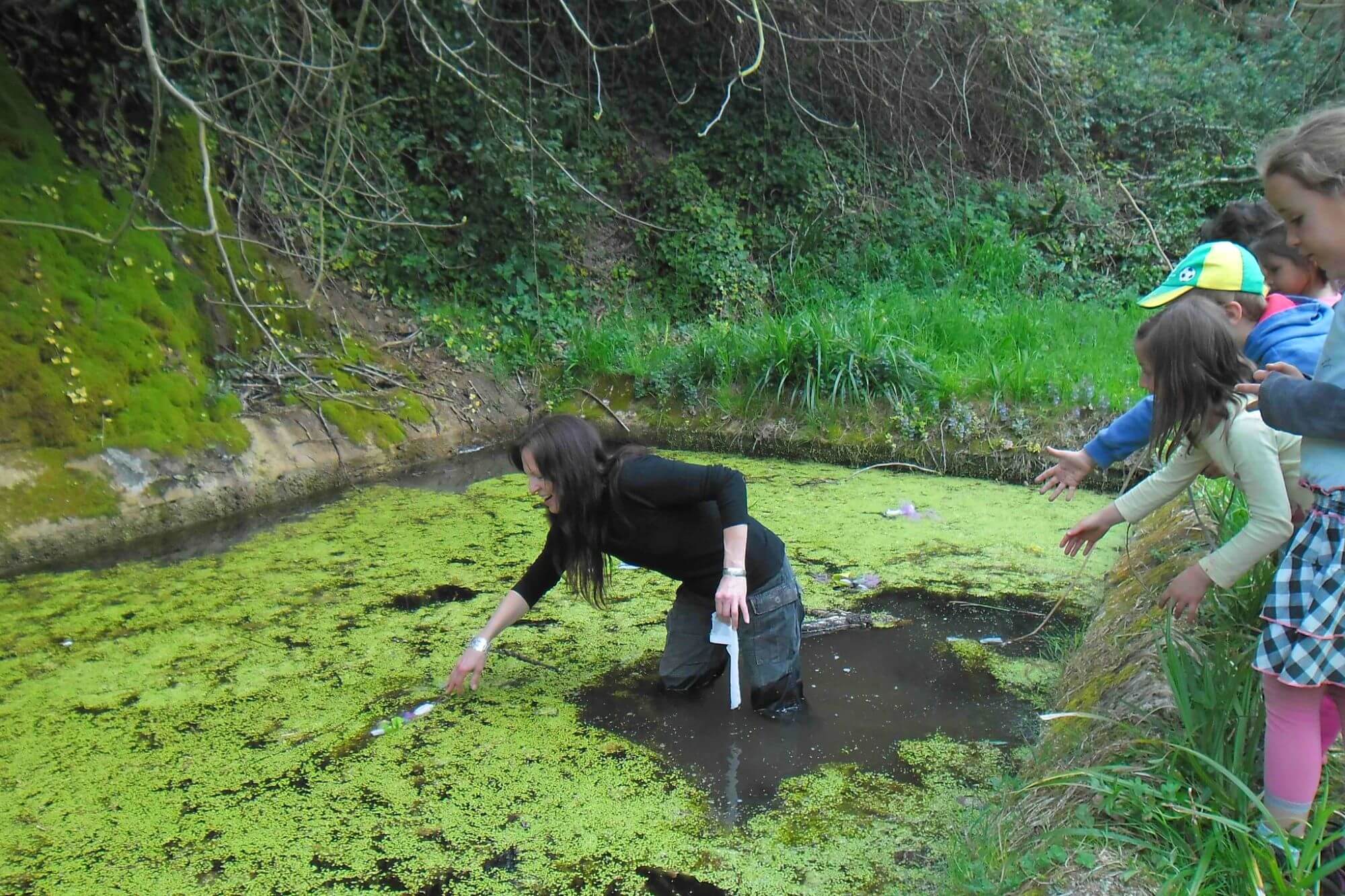 Photographie de l'artiste Deborah Bowman, lors d'une intervention pédagogique. Elle a les pieds dans l'eau d'un lavoir, au milieu des lentilles d'eau. Des enfants observent sur le bord du lavoir.