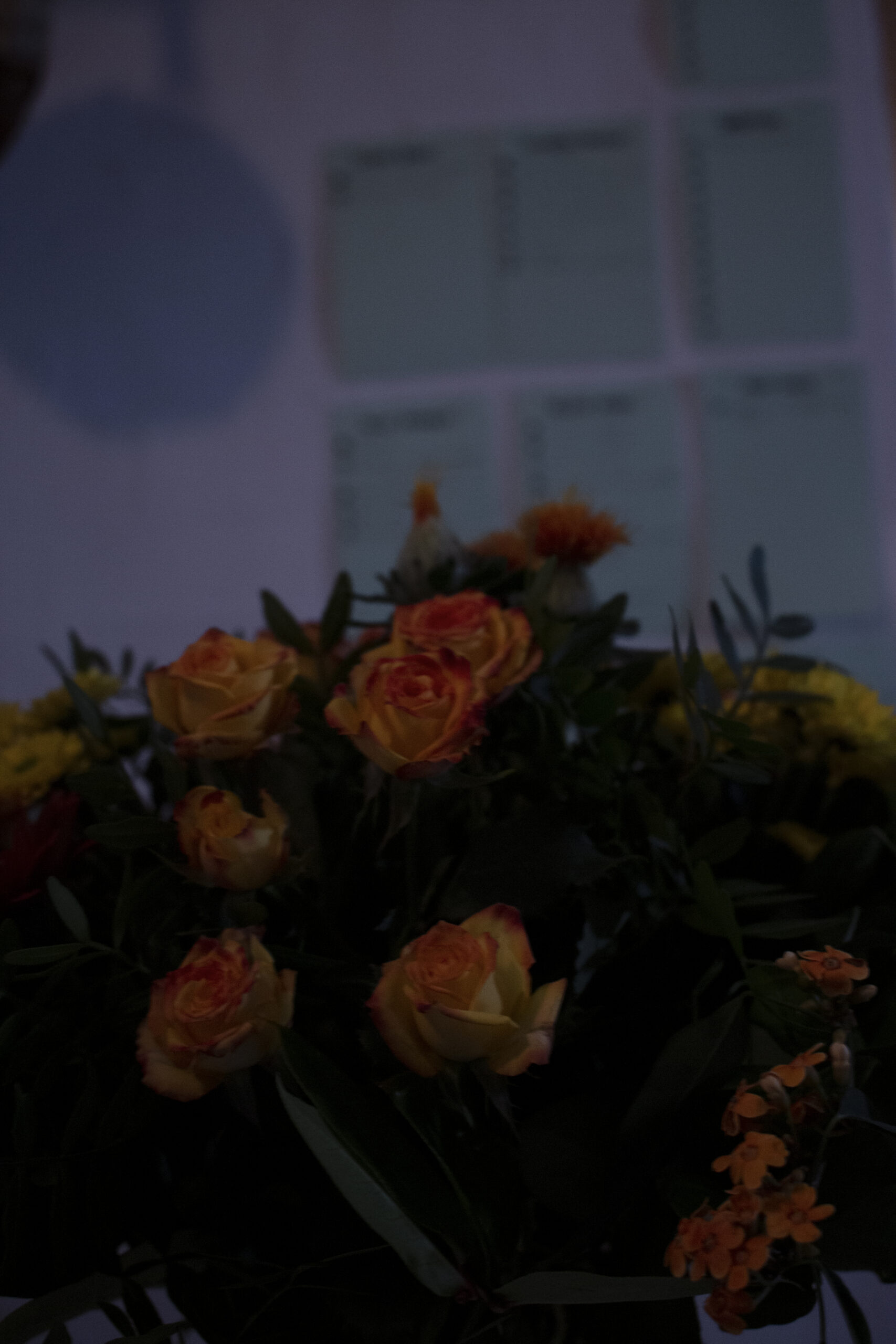 Au premier plan, un bouquet de fleurs avec en particulier des tons orangers. A l'arrière plan, des fiches de travail accroché au mur. L'arrière plan est flou.