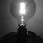 Une lampe de chevet avec une très grosse ampoule poussiéreuse est allumée. L'image est en noir et blanc.