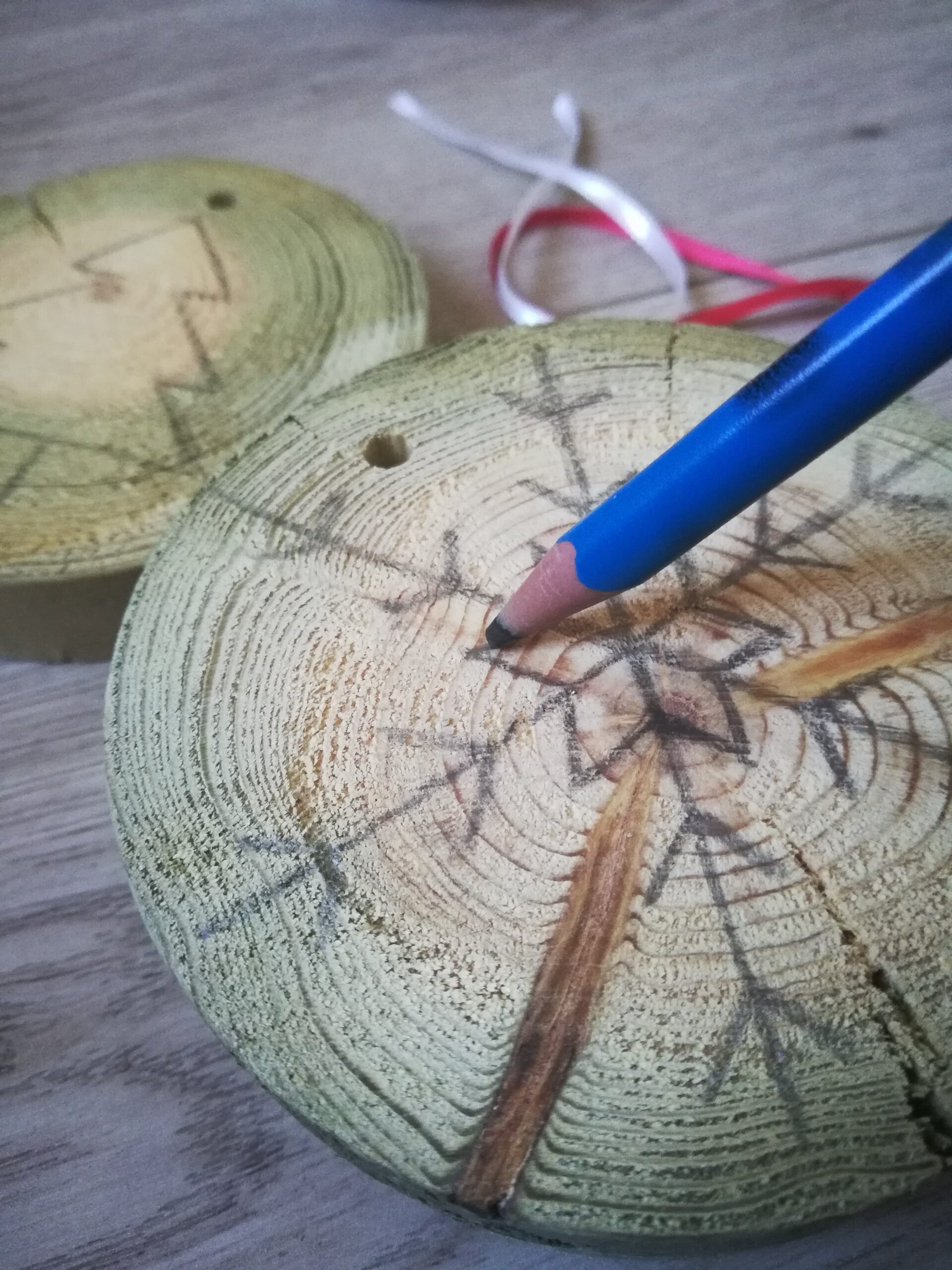 Sur une rondelle de bois, un flocon de neige se dessine. Un crayon de papier a sa pointe posée sur le bois, quelqu'un que nous ne voyons pas le tient et dessine avec.