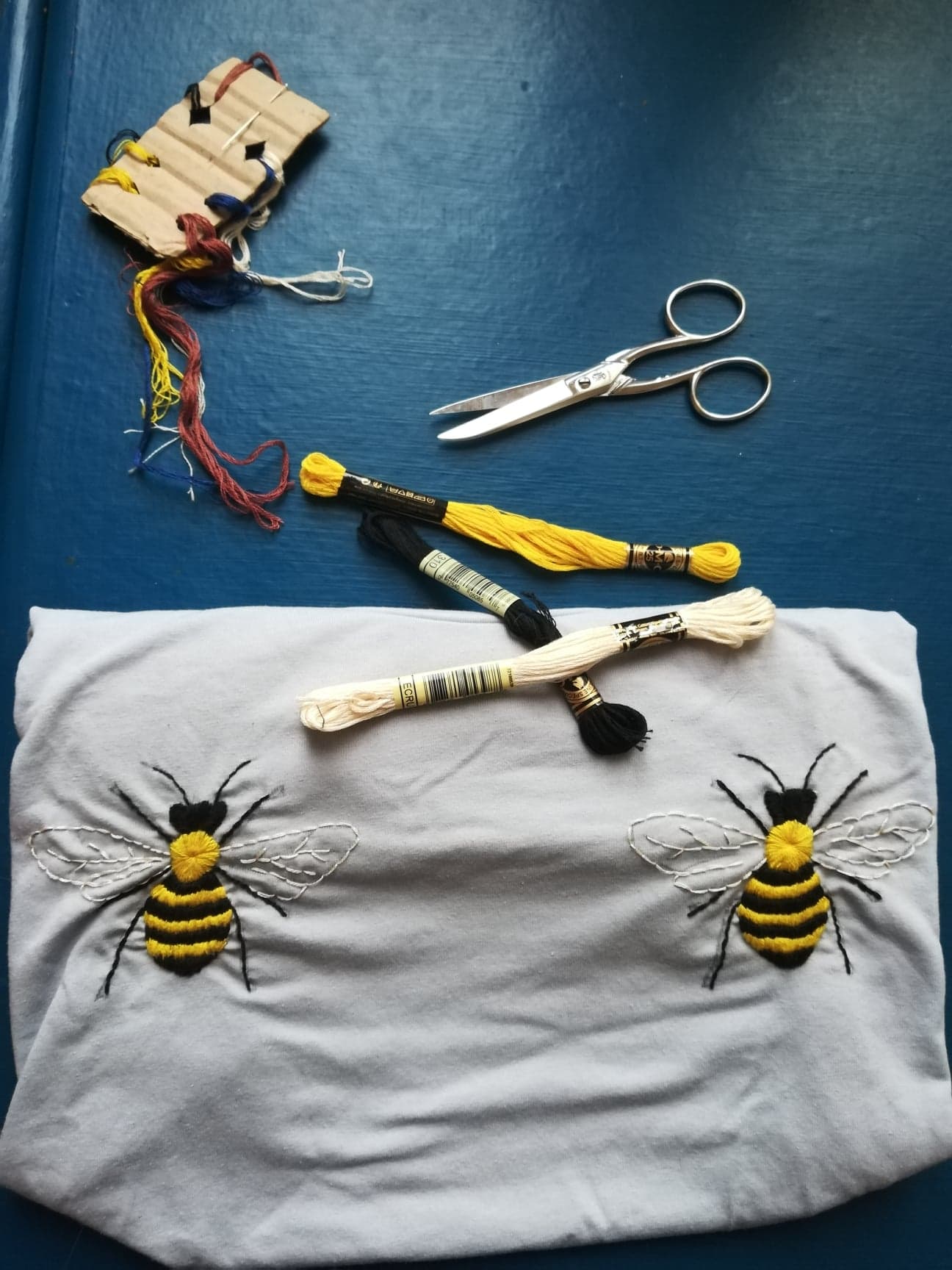 Sur un table un kit de broderie est posé. Une paire de ciseaux, des fils jaunes, blancs et noirs. Deux abeilles sont brodées symétriquement sur un T-shirt mais elles ne sont pas totalement terminées, les ailes sont encore à remplir.