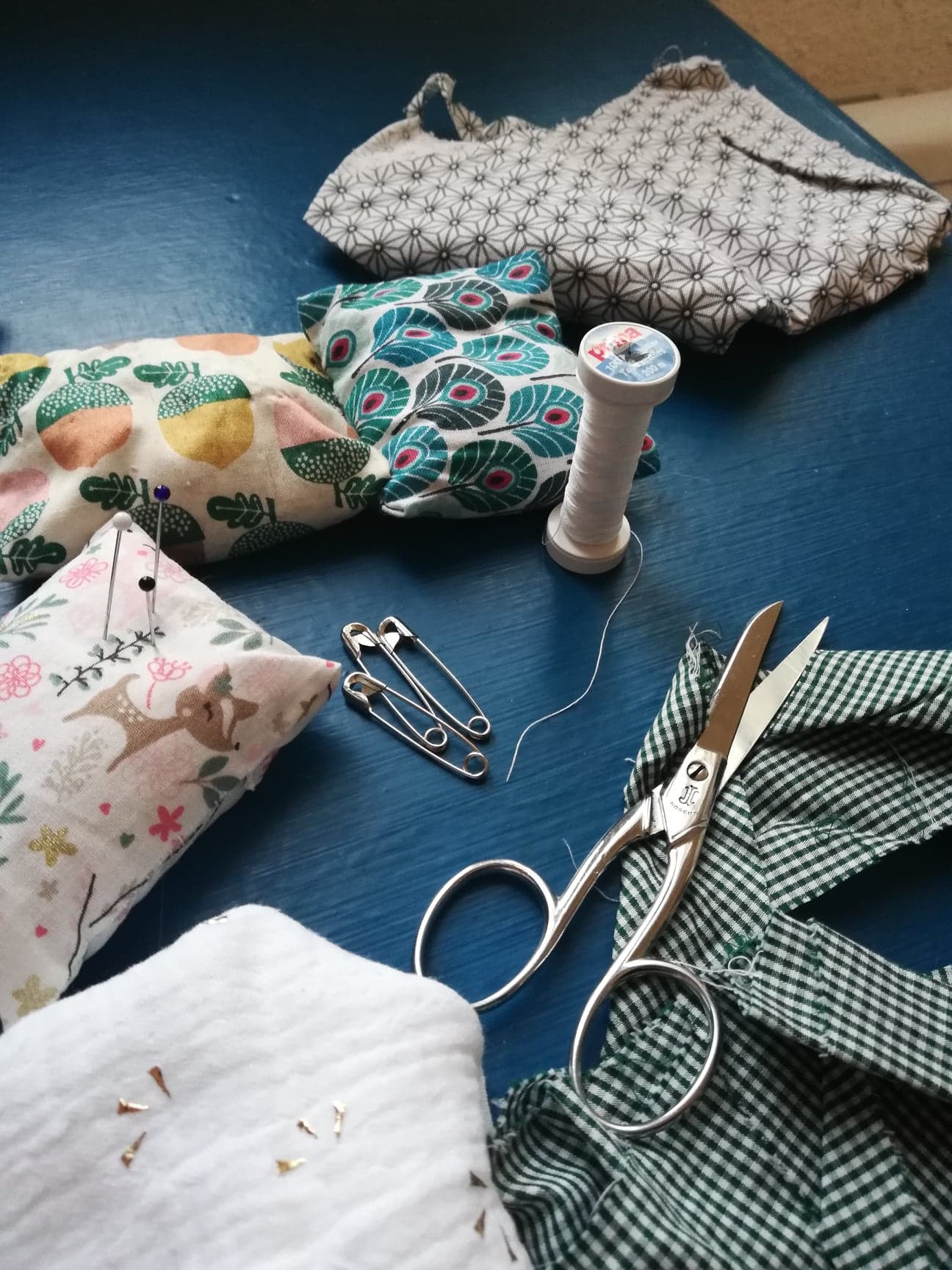 Tout un kit de couture est posé sur une table. Une paire de ciseaux, du fil blanc, des épingles à nourrice et plusieurs tissus colorés et à motifs (des petits cerfs, des carreaux, des glands).