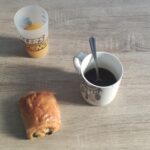 Gros plan sur un petit déjeuner. Il est posé sur une table marron clair. En haut, du jus d'orange, au centre, un café, une cuillère dans une tasse blanche. En bas, une chocolatine.