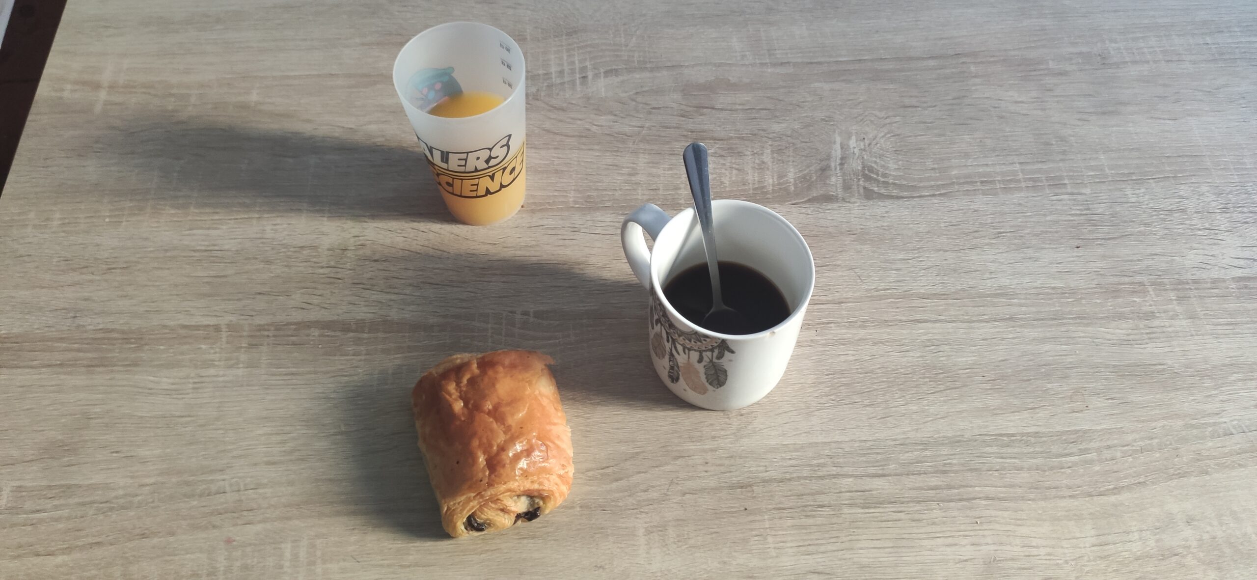 Gros plan sur un petit déjeuner. Il est posé sur une table marron clair. En haut, du jus d'orange, au centre, un café, une cuillère dans une tasse blanche. En bas, une chocolatine.