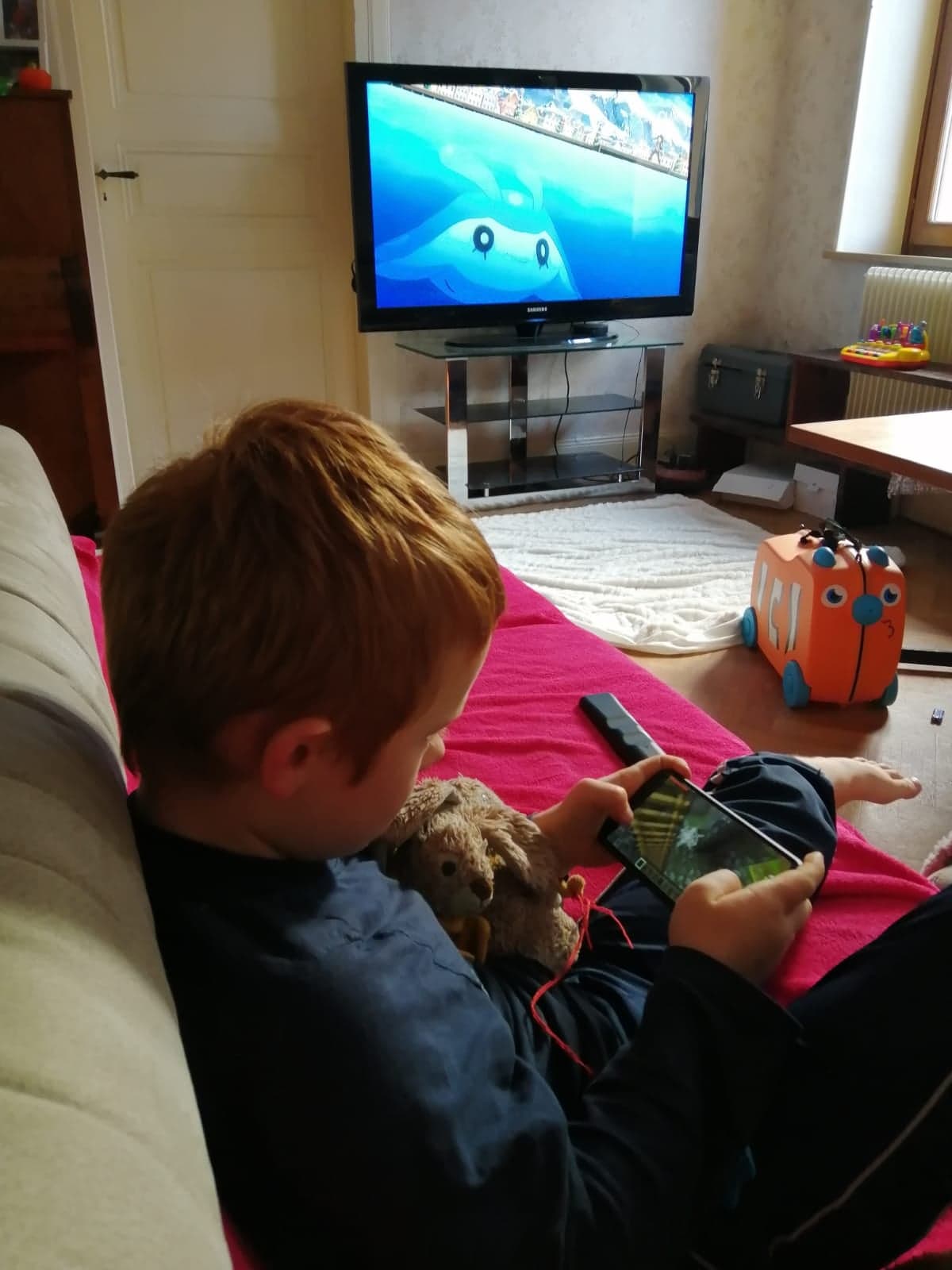 Sur cette photo on peut voir en premier plan, un petit garçon avec son doudou dans les bras, en train de jouer à un jeu d'action sur un téléphone portable. En arrière-plan, la télévision est allumée et joue un film d'animation qui ne semble pas intéresser le garçon.