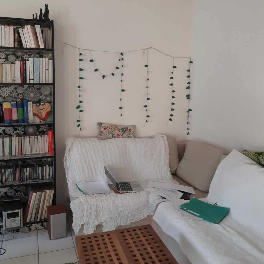 Le bureau de la chercheuse est en fait son canapé, entouré de guirlandes et de couvertures. Sur le canapé, on voit un ordinateur et plusieurs feuilles et pochettes. Sur le côté, on voit aussi une bibliothèque remplie de livres.