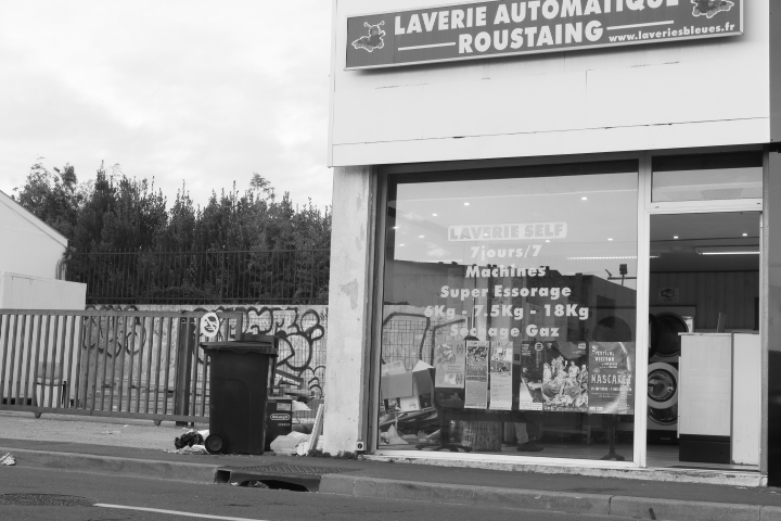 L'extérieur d'une laverie. Un panneau sur le haut du bâtiment indique "Laverie automatique roustaing". Sur la gauche de l’image il y a des poubelles ou une décharge, juste à coté de la laverie. L'image est en noir et blanc.