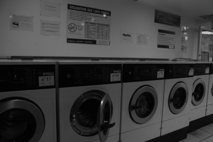 L'intérieur d'une laverie avec plusieurs machine à laver dont une ouverte et trois fermées. Il y a aussi un panneau d'instructions pour l'utilisation des machines. L'image est en noir et blanc.