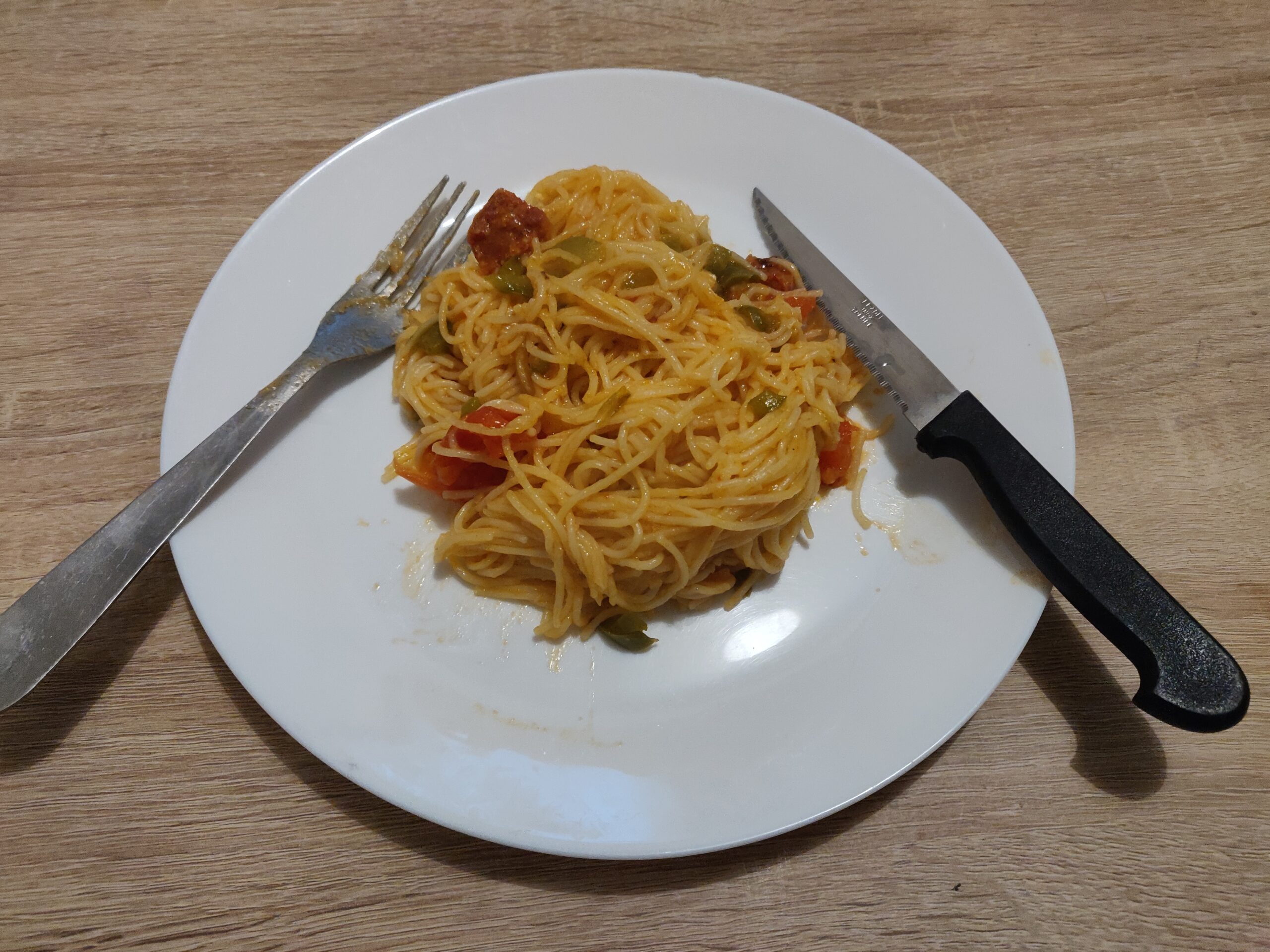 Au centre de l'image une assiette blanche, remplie de pâtes aux poivrons. Une fourchette est posée sur la gauche de la photo, un couteau, sur la droite.