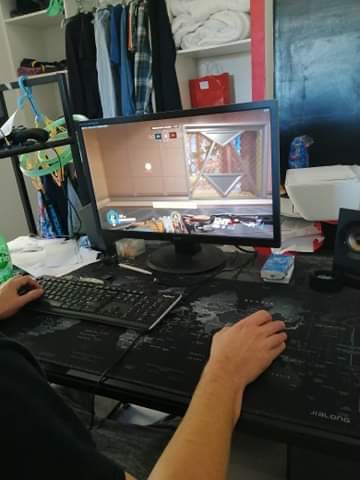 On voit les deux mains d'un jeune homme en train de jouer à un jeu vidéo. Une main tenant sa souris et l'autre active sur son clavier. Son écran d'ordinateur de bureau montre une arme qu'il doit guider pour attaquer ou se défende.