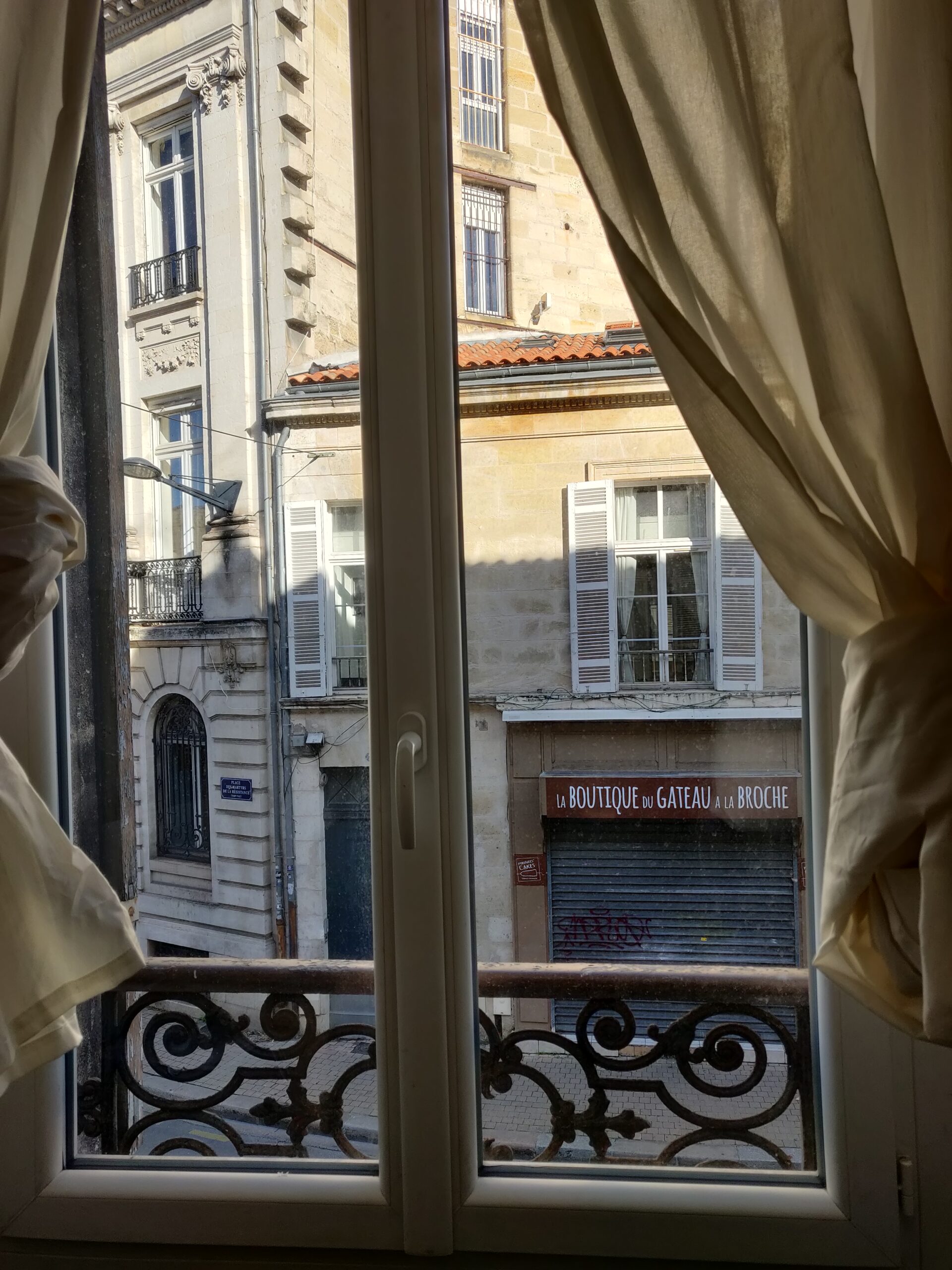 Vue d'une fenêtre. Les rideaux de couleur blanc cassé sont noués. On aperçoit les bâtiments d'en face. Particulièrement "la boutique du gâteau à la broche", rideau fermé, et orné d'un tag rouge.