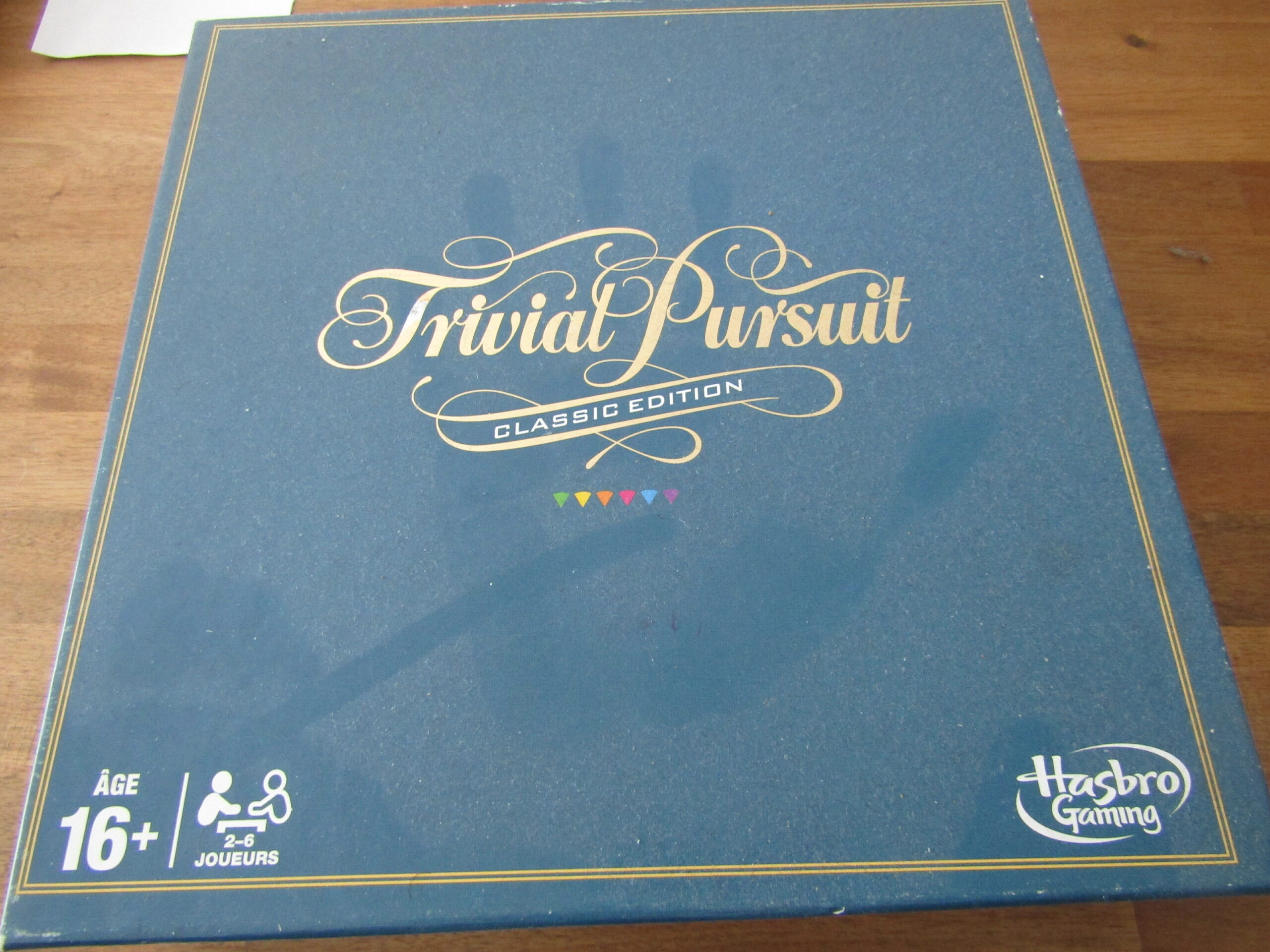 Un jeu de société Trivial Pursuit poussiéreux avec une trace de main sur le carton. Le carton est de couleur unie avec le nom du jeu par-dessus, au centre de la boîte.