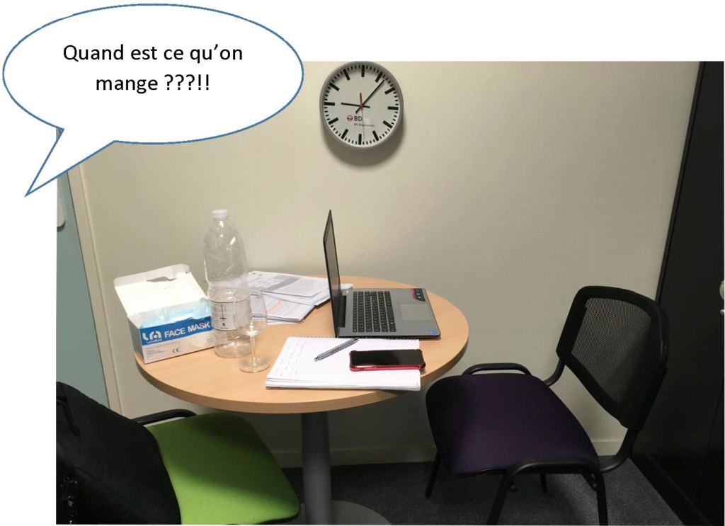 Sur un bureau rond sont disposés un ordinateur portable, une bouteille d'eau, un téléphone portable, un cahier et un stylo ainsi qu'une boîte de masques chirurgicaux. 