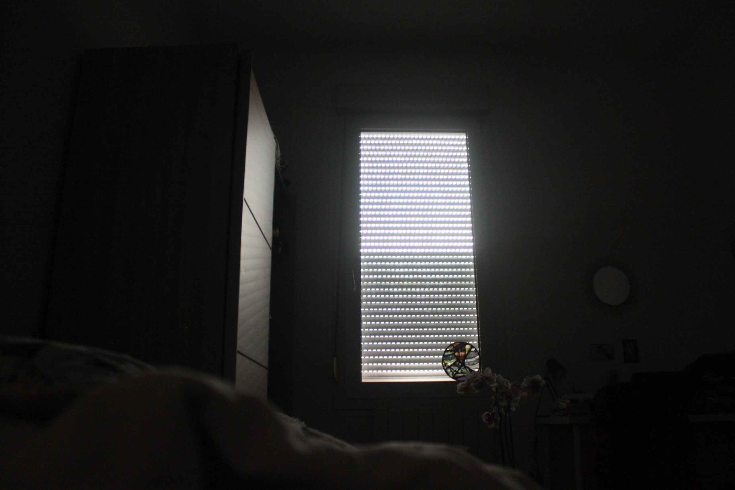 La vue depuis le lit de Nina. Une simple fenêtre avec les volets roulants fermés. La lumière passe à travers.