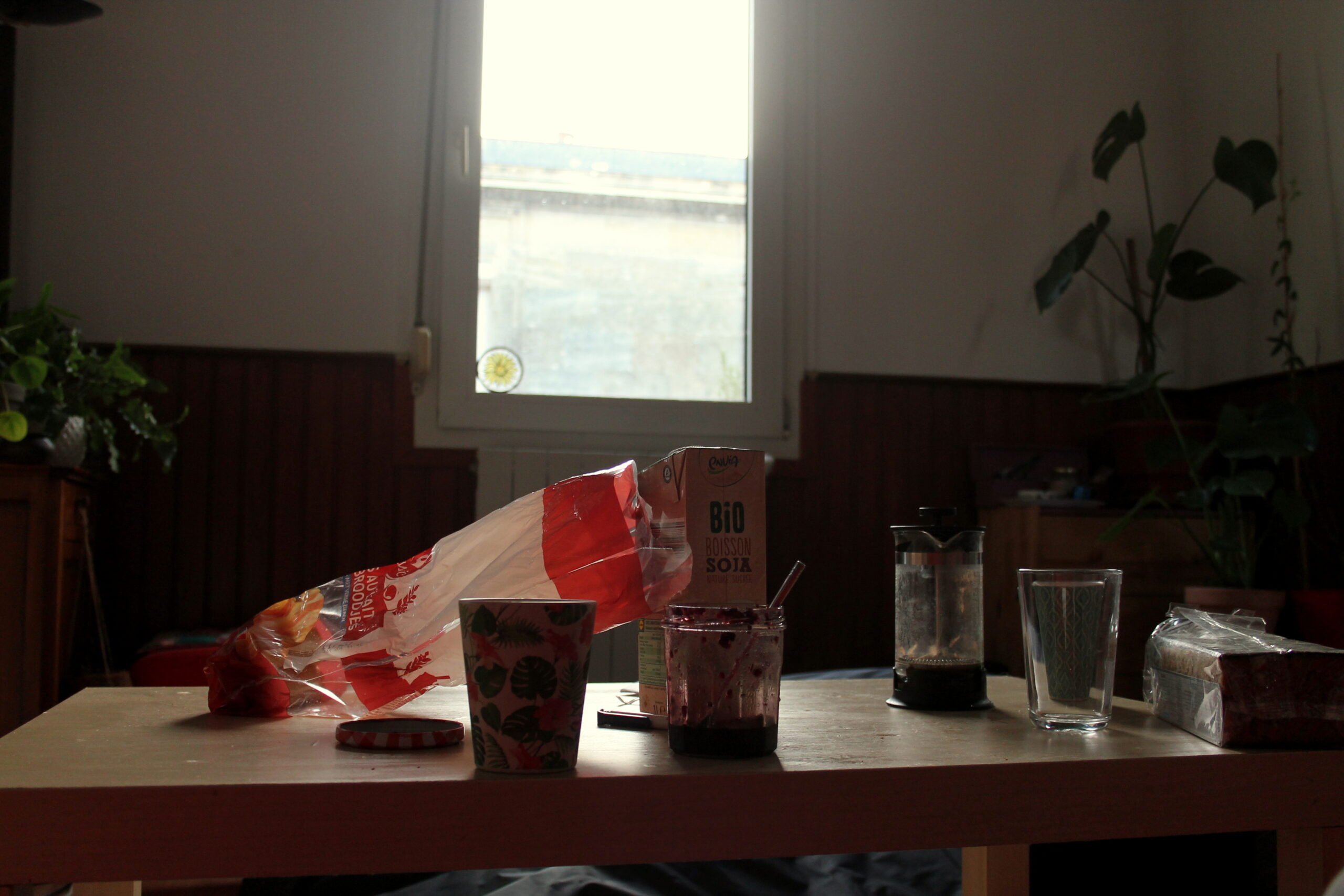 Nina prend son petit-déjeuner. Sur la table il y a du café de la confiture, du lait de soja et des petits pains au lait. En fond, une fenêtre avec les volets ouverts.