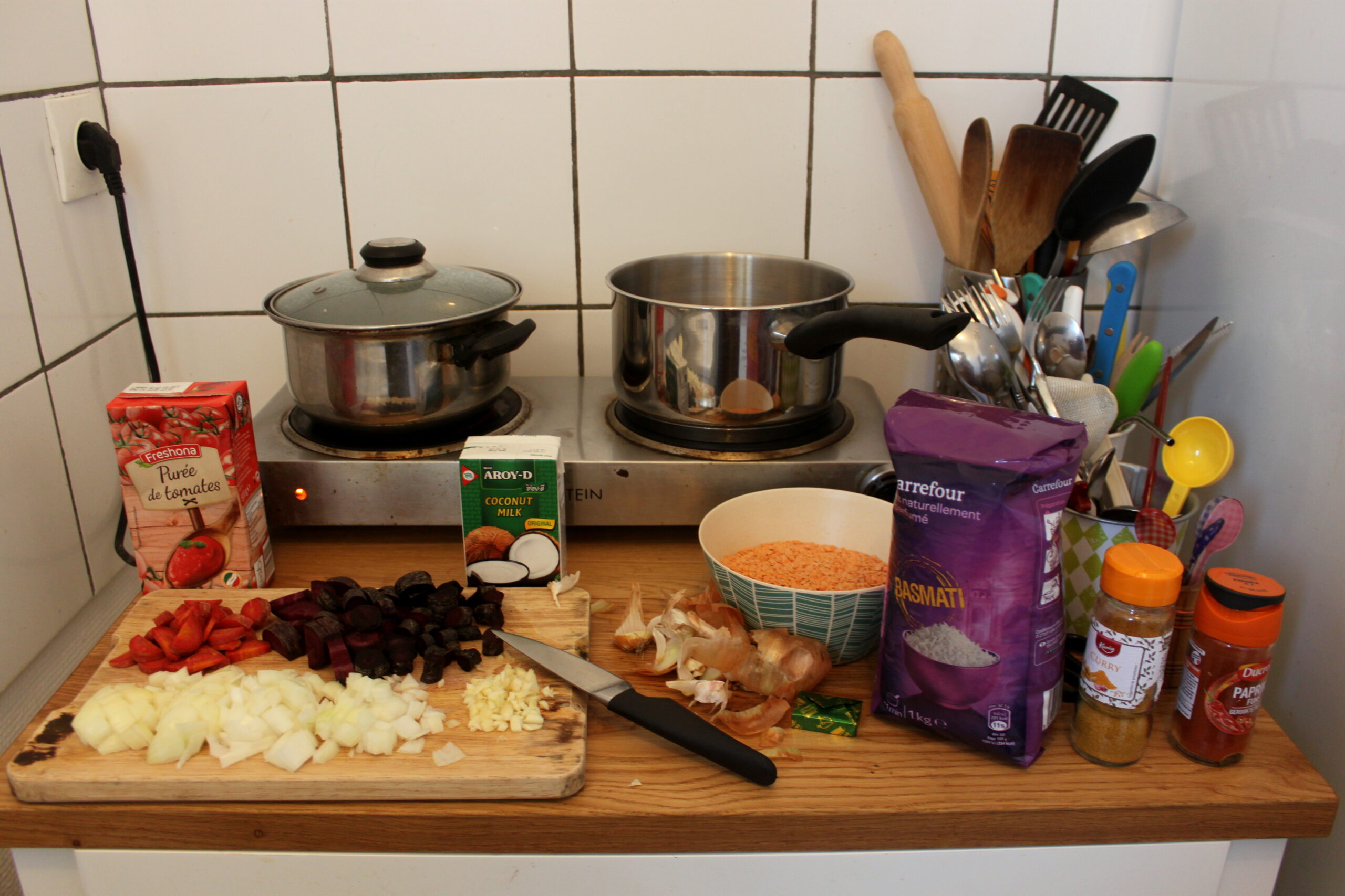 La cuisine de Nina. Sur un petit meuble il y a tout le nécessaire pour faire un curry de lentilles corail. Deux petites plaques électriques sont au fond avec deux casseroles posées dessus.