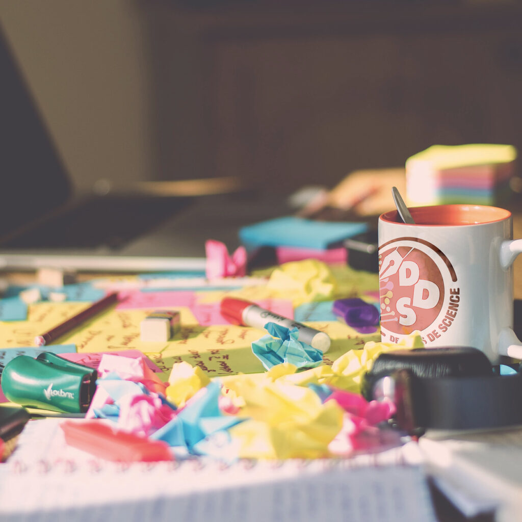 Image avec tasse dans un casque audio à droite, des papiers multicolores au centre et un pc ouvert en arrière plan flouté à gauche